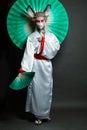 Cheerful woman in animal mask and white kimono standing with green fan on black background, full length portrait. Halloween,