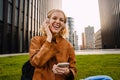 Cheerful woman listening music with headphones and mobile phone while sitting on grass near modern office buildings Royalty Free Stock Photo