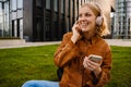 Cheerful woman listening music with headphones and mobile phone while sitting near modern office buildings Royalty Free Stock Photo