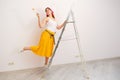 Cheerful woman painter with a brush during the repair. Girl decorator smiling standing on a ladder against the wall, copy space
