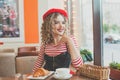 Cheerful woman in red beret smiling and drinking coffee in cafe