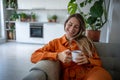 Cheerful woman with smile recalls happy moment from past while sits with cup of tea on sofa at home. Royalty Free Stock Photo