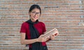 Cheerful woman student wearing glasses standing and holding book with brick wall as background. Education concept Royalty Free Stock Photo