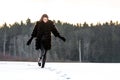 A cheerful girl in a warm fur coat walks across the open field leaving footpath in the snow