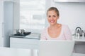 Woman sitting at kitchen counter using silver laptop, wearing pink top, stainless pots visible Royalty Free Stock Photo