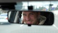 Cheerful adult Caucasian male driving a car in city looking and smiling in rearview mirror. Happy man face reflected in Royalty Free Stock Photo