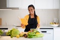 Cheerful young african american female in apron preparing salad and reading recipe book Royalty Free Stock Photo