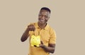 Cheerful young African American man tossing money into piggy bank making savings stands in studio Royalty Free Stock Photo