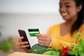 Cheerful young african american woman with paper bag with organic vegetables uses smartphone and credit card Royalty Free Stock Photo