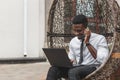 Cheerful young African man in formal wear talking on the mobile phone and working on laptop while sitting on egg chair Royalty Free Stock Photo