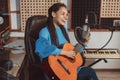 Cheerful young African woman, singer guitarist playing guitar and singing song into microphone in sound recording studio
