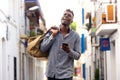 Cheerful young afro american guy holding handbag walking outside with mobile phone Royalty Free Stock Photo