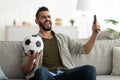 Cheerful young Arab guy watching soccer game on TV, holding ball and remote control, sitting on couch at home Royalty Free Stock Photo
