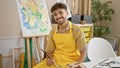 Cheerful young arabian man, an artistic prodigy, laughing delightedly, holding paintbrush and palette, sitting in an art studio, Royalty Free Stock Photo