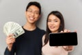 Cheerful young asian loving couple holding money and showing display of mobile phone. Royalty Free Stock Photo