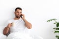 Cheerful young attractive guy with beard speaks by phone, drink coffee at free time in minimalist bedroom interior Royalty Free Stock Photo