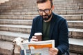 Cheerful young bearded man sitting outdoors on steps reading newspaper Royalty Free Stock Photo