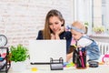 Cheerful young beautiful businesswoman looking at laptop while sitting at her working place with her little daughter Royalty Free Stock Photo