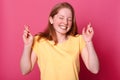 Cheerful young beautiful girl with crossed fingers, makes wish, smiles broadly, wears casual yellow t shirt, models over pink wall Royalty Free Stock Photo