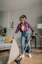 Cheerful young black female in wireless headphones with vacuum cleaner dusts floor, enjoys music in living room