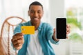 Cheerful young black male shopaholic in casual showing credit card and smartphone with blank screen