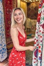 Cheerful young blonde woman in front of carpet and fabric market in wavy red summer dress. Young beautiful woman entering shop. Royalty Free Stock Photo