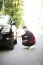 Cheerful young indian man has problems with the wheel of his car. He is kneeing and looking at it with seriousness