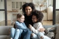 Cheerful African mother sitting on couch embraces multiracial daughters