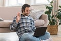 Cheerful young caucasian guy sits floor, waving hand at computer webcam, has video call in room interior