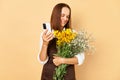 Cheerful young caucasian woman florist wearing brown apron holding flowers isolated over beige background using mobile phone Royalty Free Stock Photo