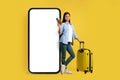 Cheerful young chinese woman in casual with suitcase passport and airtickets near huge smartphone with blank screen Royalty Free Stock Photo