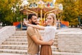 Cheerful young couple in love standing hugging smiling looking away in amusement park on background of carousel on Royalty Free Stock Photo