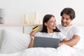 Cheerful young european male with stubble hugs woman sitting on bed use credit card and laptop for online shopping