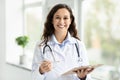 Cheerful young european woman doctor posing by window, holding clipboard Royalty Free Stock Photo