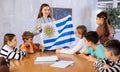 Cheerful young female teacher showing flag of Uruguay to schoolchildren preteens during history lesson in audience