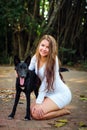 Cheerful young girl on walk in the park with her four-legged friend. Pretty woman in short dress and black dog playing outdoors.