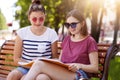 Cheerful young girls spend time together in hot sunny day in the park. Beauties look through youth magazine while sitting on Royalty Free Stock Photo