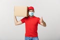 Cheerful young handsome delivery man holding a cardboard box and showing his thumb up while standing against grey Royalty Free Stock Photo
