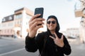 Cheerful young hipster man in hoodie in sunglasses in trendy baseball cap is smiling and showing peace sign making selfie on phone Royalty Free Stock Photo