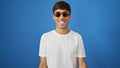 Cheerful young hispanic man wearing sunglasses, exuding confidence and positivity, standing against an isolated blue background Royalty Free Stock Photo