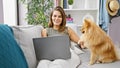 Cheerful, young hispanic woman sitting on the sofa, using laptop with her playful dog, enjoying online technology in the comfort Royalty Free Stock Photo