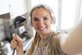 Cheerful young housewife in apron and earphones with soup ladle taking selfie photo in cozy kitchen