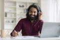 Cheerful young indian man working from home office, using laptop and headset Royalty Free Stock Photo