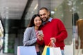 Cheerful young interracial couple with gift bags and mobile phone ordering goods online near shopping mall Royalty Free Stock Photo