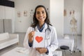 Cheerful young Latin cardiologist holding greeting card with red heart