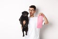 Cheerful young man holding black pug and pink dog poop bag, standing over white background