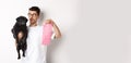 Cheerful young man holding black pug and pink dog poop bag, standing over white background