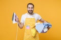 Cheerful young man househusband in apron hold basket with clean clothes, iron doing housework isolated on yellow