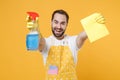 Cheerful young man househusband in apron rubber gloves hold spray with washing cleanser, cleaning rag while doing
