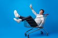 Cheerful young man sitting in shopping cart on blue background Royalty Free Stock Photo